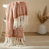 Pink and white fringed throw blanket draped over a chair in a room with a vase and plant.