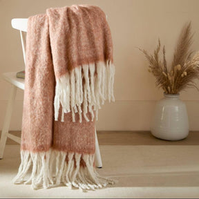 Pink and white fringed throw blanket draped over a chair in a room with a vase and plant.